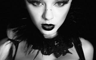 Woman in dramatic black and white makeup with a striped outfit and feathered collar, posing against a dark background.