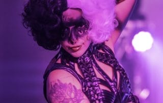 Performer with half black, half purple hair and dramatic makeup poses under purple lights at the Halloween event.
