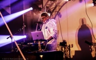 DJ performing in a white outfit with headphones at a Denver Halloween event, illuminated by colorful stage lights.