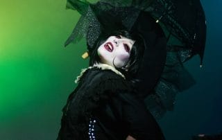Performer in dramatic black costume and makeup, striking a pose with a black umbrella under colorful stage lights.