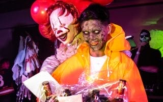 A clown holding red balloons stands behind a zombie in an orange raincoat, both smiling at a Halloween event.