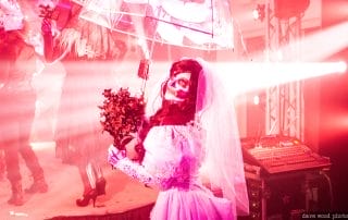 A bride in a skeleton costume holds a bouquet, illuminated by pink lights at a Halloween event.