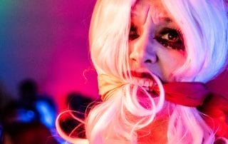 A person with a white wig and dramatic makeup, looking intensely at the camera under colorful lights.