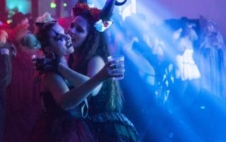 Two women in elaborate costumes share a joyful moment on the dance floor, surrounded by colorful lights at the event.