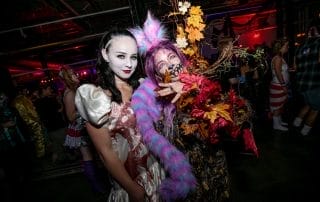 Two women in creative costumes pose together, one in a blood-splattered gown and the other adorned with colorful foliage.