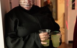 Person in a striking costume with pins in their head, holding two drinks at the Paranormal Palace event.