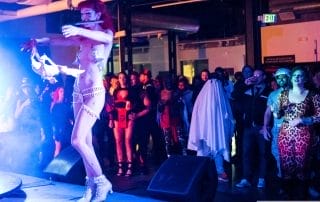 A performer in a sparkling costume dances on stage while an engaged crowd watches at the Halloween event in Denver.