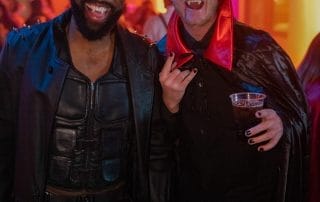 Two men in Halloween costumes smiling at the camera, one in a leather outfit and the other as a vampire, at a party.