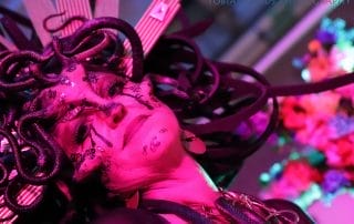 A performer with a snake-themed headdress and intricate face paint under vibrant pink lighting at the Halloween event.