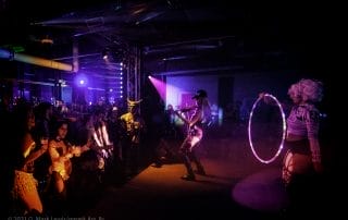 A performer with a microphone entertains the crowd while another hula hoops in colorful lights at the Halloween event.