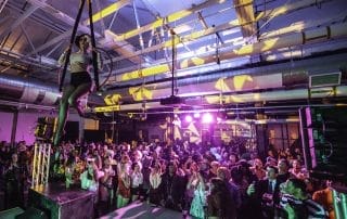 Aerial performer swinging above a lively crowd at the Paranormal Palace event in Denver. Colorful lights illuminate the scene.