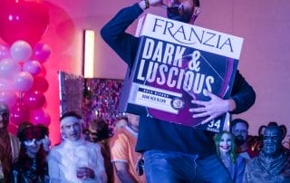 A man in a black shirt drinks from a cup while holding a large Franzia wine box at a Halloween event.