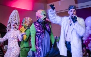 Costume contest participants showcasing creative outfits on stage, including an alien and a character in a white fur coat.