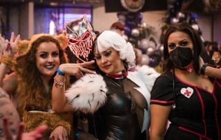 Three women in creative costumes pose together, showcasing a lion, a character with white hair, and a nurse outfit.